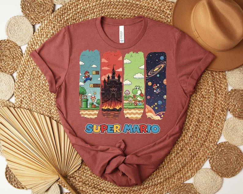 May include: A maroon t-shirt featuring a Super Mario graphic. The design includes four vertical panels with scenes from the game, including Mario, a castle, Yoshi, and a space scene. The words "SUPER MARIO" are printed in a colorful font.