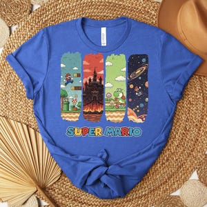 May include: A royal blue t-shirt featuring a colorful graphic of Super Mario characters and scenes. The design includes images of Mario, a castle, Yoshi, and a space scene. The words "SUPER MARIO" are printed in a colorful font.