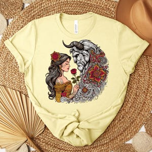 May include: Light yellow t-shirt featuring a detailed graphic of a woman and a beast, holding a rose. The design includes floral and ornamental elements in red, gold, and black. The shirt is made of a soft material.