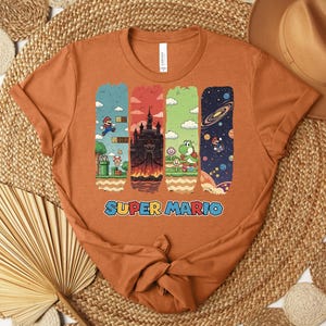 May include: An orange t-shirt featuring a Super Mario design. The graphic includes four vertical panels with scenes from the game, including Mario, a castle, Yoshi, and a space scene. The words "SUPER MARIO" are printed in blue.