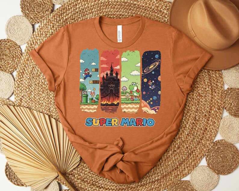 May include: An orange t-shirt featuring a Super Mario design. The graphic includes four vertical panels with scenes from the game, including Mario, a castle, Yoshi, and a space scene. The words "SUPER MARIO" are printed in blue.