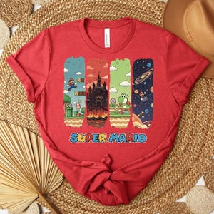 May include: A red t-shirt featuring a Super Mario design. The graphic includes four vertical rectangular panels with scenes from the game, including Mario, a castle, Yoshi, and a space scene. The words "SUPER MARIO" are printed in blue.