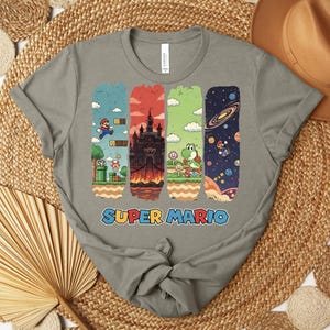 May include: A gray t-shirt featuring a colorful Super Mario graphic. The design includes iconic scenes from the game, with Mario jumping, a castle, Yoshi, and a space scene. The words "SUPER MARIO" are printed in blue and red.