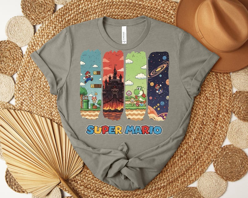 May include: A gray t-shirt featuring a colorful Super Mario graphic. The design includes iconic scenes from the game, with Mario jumping, a castle, Yoshi, and a space scene. The words "SUPER MARIO" are printed in blue and red.