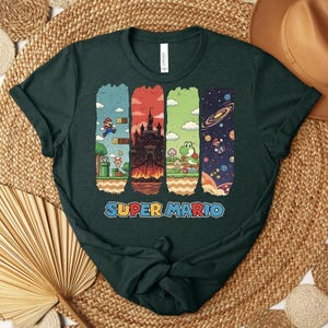 May include: Dark green t-shirt featuring a colorful Super Mario graphic. The design includes scenes from the game, such as Mario jumping, a castle, Yoshi, and a space scene. The words "SUPER MARIO" are printed in blue and orange.
