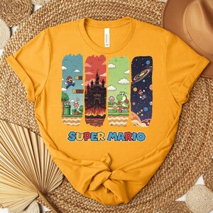 May include: Mustard yellow t-shirt featuring a Super Mario graphic. The graphic includes four vertical panels with scenes from the game, including Mario, a castle, Yoshi, and a space scene. The words "SUPER MARIO" are printed in blue.