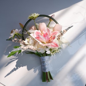 Puede incluir: Un ramo de novia con un gran lirio rosa y blanco como punto focal, rodeado de rosas blancas, tulipanes rosas y otras flores delicadas. Los tallos están envueltos en una cinta blanca, creando un arreglo elegante.