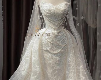 Op de afbeelding: Een witte strapless kanten trouwjurk met een hartvormige halslijn, ingewikkeld bloemenborduurwerk en gedrapeerde parelsnoeren over het lijfje. De japon bevat een bijpassende doorschijnende sluier versierd met parels en de tekst TIEM VAY CUOI TVC870.