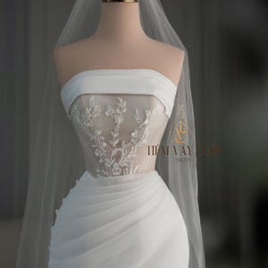 May include: A strapless white wedding dress with a sheer, embroidered bodice and a draped skirt. A gold crown and a long veil are on the mannequin. The text "TIEM VAY CUOI" is visible on the right side of the dress.