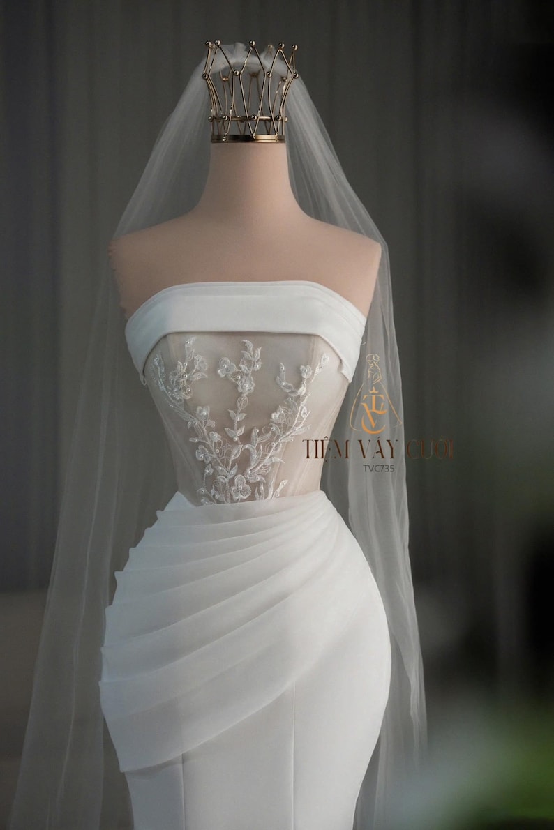 May include: A strapless white wedding dress with a sheer, embroidered bodice and a draped skirt. A gold crown and a long veil are on the mannequin. The text "TIEM VAY CUOI" is visible on the right side of the dress.