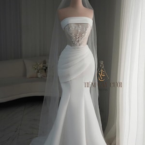 May include: A white strapless wedding dress on a mannequin. The bodice features delicate lace detailing, and the skirt flows into a mermaid silhouette. A sheer veil and a gold crown complete the bridal look. The text "TIỆM V&Aacute;Y CƯỚI" is visible.