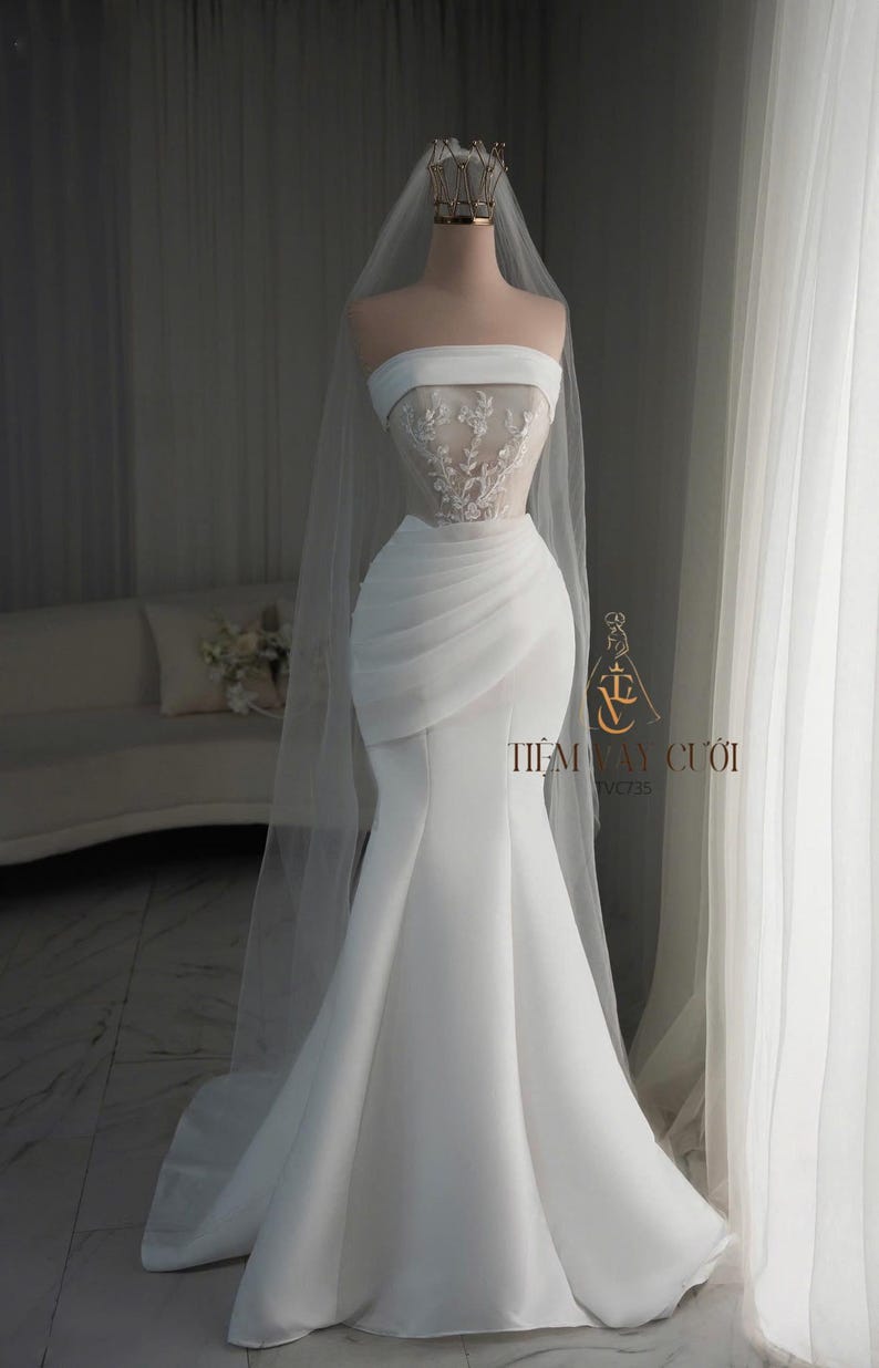 May include: A white strapless wedding dress on a mannequin. The bodice features delicate lace detailing, and the skirt flows into a mermaid silhouette. A sheer veil and a gold crown complete the bridal look. The text "TIỆM V&Aacute;Y CƯỚI" is visible.