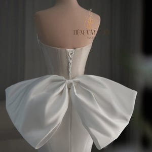 May include: A white wedding dress on a mannequin, featuring a large bow detail at the back. The dress has a corset-style bodice and a lace-up back. A gold crown sits atop the mannequin's head. The text "TIEM VAY CUOI" is visible.