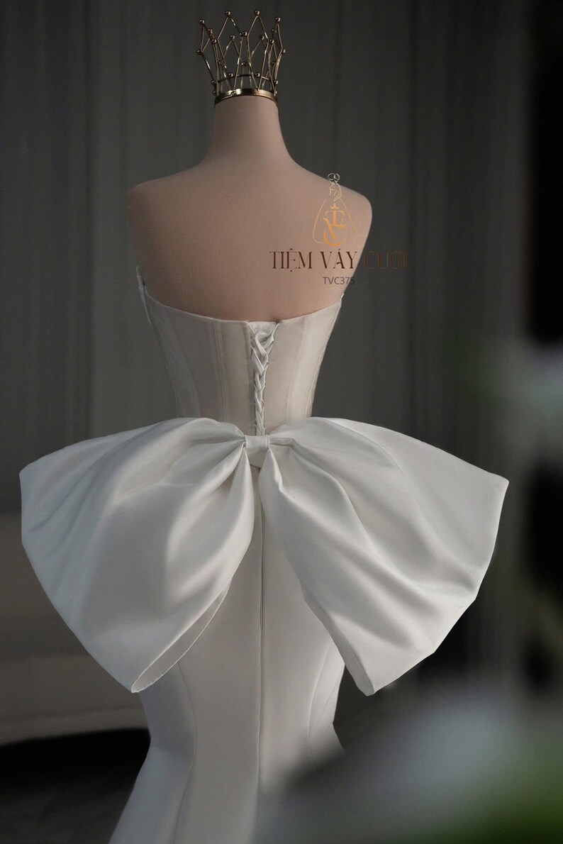May include: A white wedding dress on a mannequin, featuring a large bow detail at the back. The dress has a corset-style bodice and a lace-up back. A gold crown sits atop the mannequin's head. The text "TIEM VAY CUOI" is visible.