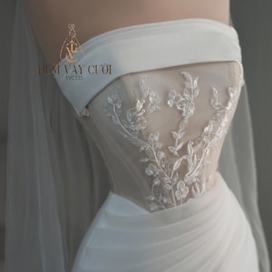 May include: A strapless white wedding dress with delicate floral embroidery on the bodice. The dress features a draped skirt and a sheer veil. The logo "THIẾT VAY CƯỚI" is visible.