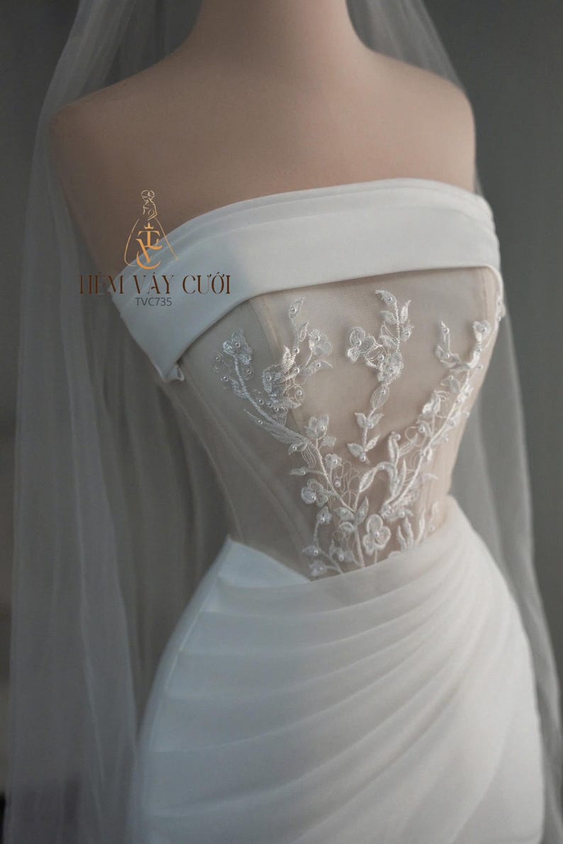 May include: A strapless white wedding dress with delicate floral embroidery on the bodice. The dress features a draped skirt and a sheer veil. The logo "THIẾT VAY CƯỚI" is visible.
