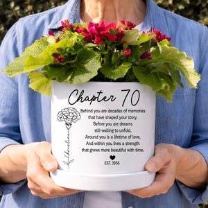May include: A white ceramic flower pot featuring the text Chapter 70, a floral illustration, and a poem about memories and dreams. The pot includes the name Christina, a small heart icon, and the text EST. 1956, filled with a vibrant red flowering plant.