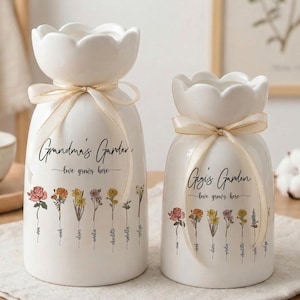 May include: Two white ceramic vases with a floral design and the text "Grandma's Garden" and "Gigi's Garden". Each vase has a scalloped top, a cream-colored ribbon, and a row of colorful flowers. The words "love grows here" are also written on the vases.