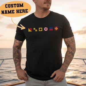 Può includere: T-shirt nero girocollo con una fila di bandiere nautiche colorate ricamate sul petto. La maglietta ha maniche corte. Un ovale arancione con la scritta "CUSTOM NAME HERE" si trova nell'angolo in alto a sinistra.