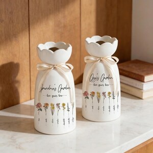 May include: Two white ceramic vases with a floral design and the text "Grandma's Garden" and "Gigi's Garden". Each vase has a scalloped top, a cream-colored ribbon, and a series of colorful flowers printed on the front.