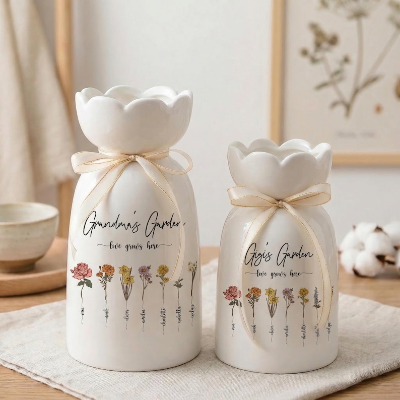 May include: Two white ceramic vases with a floral design and the text "Grandma's Garden" and "Gigi's Garden". Each vase has a scalloped top, a cream-colored ribbon, and a row of colorful flowers. The words "love grows here" are also written on the vases.