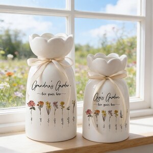 May include: Two white ceramic vases with a floral design and ribbon accents. The vases have a scalloped top and feature the text "Grandma's Garden" and "Gigi's Garden" with the phrase "love grows here." The vases are decorated with colorful flower illustrations.