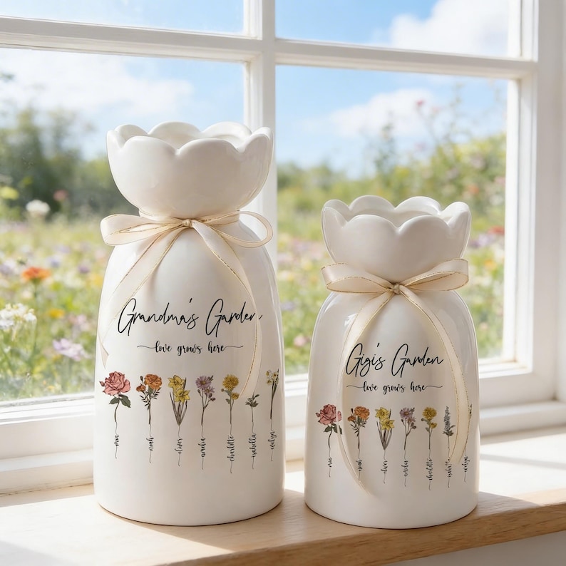 May include: Two white ceramic vases with a floral design and ribbon accents. The vases have a scalloped top and feature the text "Grandma's Garden" and "Gigi's Garden" with the phrase "love grows here." The vases are decorated with colorful flower illustrations.