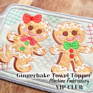 May include: A gray and white quilted kitchen towel topper with an embroidered design of two gingerbread people. The gingerbread people are smiling and have red bows and green bows. The text "Gingerbake Towel Topper Machine Embroidery VIP CLUB" is printed below the gingerbread people.