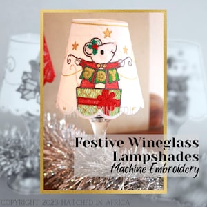 May include: White wine glass lampshade with a red and green embroidered design of a mouse wearing a festive sweater and sitting on a stack of presents. The text "Joy" is embroidered on the mouse's sweater. Festive Wineglass Lampshades Machine Embroidery.