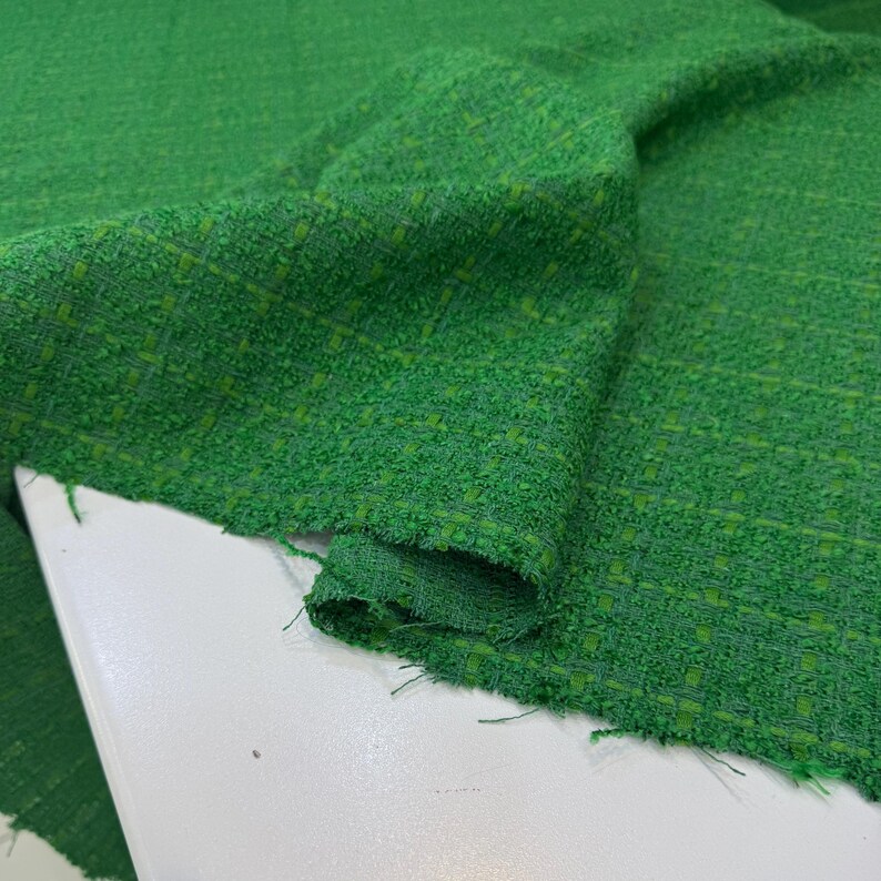 a close-up view of a green tweed fabric, possibly a piece of clothing or textile. The fabric appears to be folded or rolled, with a textured and woven appearance.