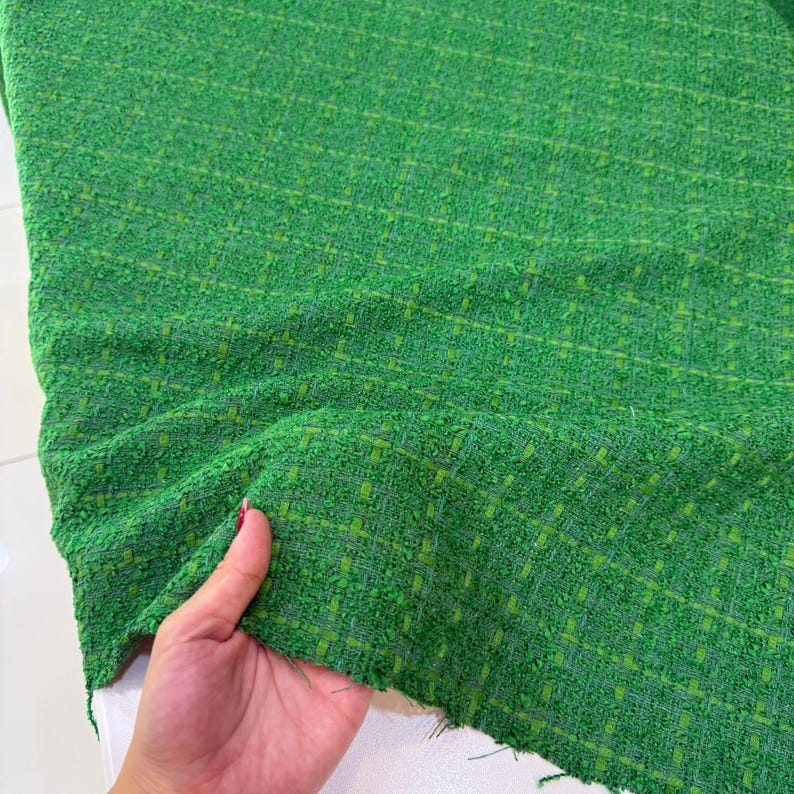 A hand is holding a piece of green tweed fabric with a pattern of small squares and lines, which appears to be a textile material.