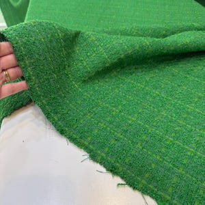 a close-up view of a green tweed fabric, with a hand holding it up to the camera. The fabric appears to be a woven material with a textured, green and white pattern.