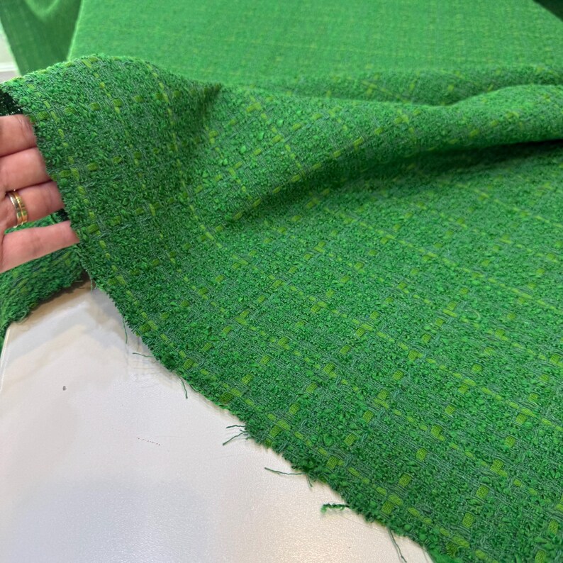 a close-up view of a green tweed fabric, with a hand holding it up to the camera. The fabric appears to be a woven material with a textured, green and white pattern.