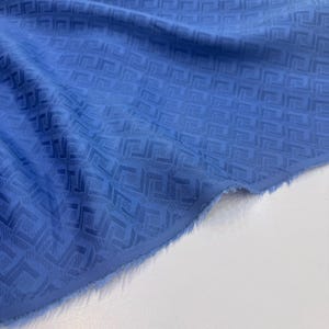 Puede incluir: Una pieza de tela azul con un patrón geométrico repetitivo de cuadrados y líneas entrelazadas. El material tiene un brillo sutil y una superficie texturizada con un borde deshilachado visible en la esquina inferior derecha.