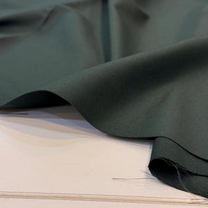 May include: Close-up of a dark green fabric with visible texture and folds. The fabric is draped over a white surface, showcasing its color and weave. The material appears to be a medium-weight textile, suitable for various sewing projects.