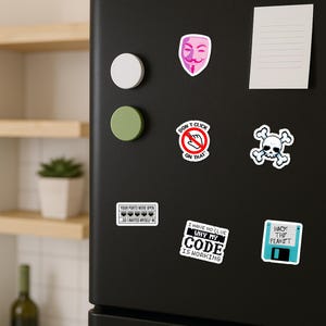 以下が含まれることがあります： 黒い冷蔵庫に貼られた、カラフルなステッカーとマグネットのコレクション。ステッカーには、ドクロとクロスボーン、ガイ・フォークスのマスク、「DON'T CLICK ON THAT!」などのテキストなど、さまざまなデザインが施されています。