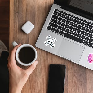 以下が含まれることがあります： 木製の表面に、スカルとクロスボーンのステッカーが付いたラップトップ、コーヒーカップ、スマートフォン、ワイヤレスイヤホンがあります。ラップトップのキーボードが表示され、画面にはテキストが表示されます。