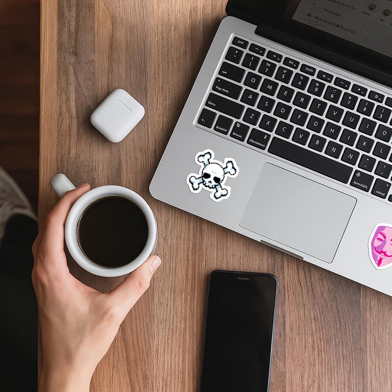 以下が含まれることがあります： 木製の表面に、スカルとクロスボーンのステッカーが付いたラップトップ、コーヒーカップ、スマートフォン、ワイヤレスイヤホンがあります。ラップトップのキーボードが表示され、画面にはテキストが表示されます。