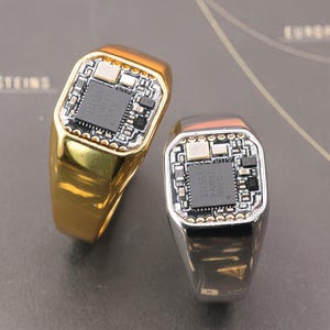 以下が含まれることがあります： 回路基板のデザインの2つの指輪。1つは金色、もう1つは銀色です。どちらも、詳細な回路基板パターンと中央のチップを備えた長方形の面があります。指輪は地図の背景に配置されています。