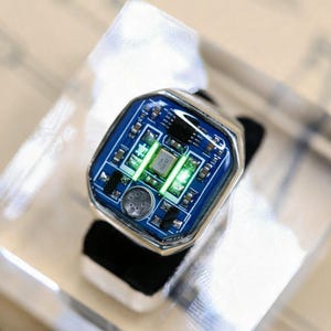 Puede incluir: Un anillo de tono plateado con una cara rectangular y una incrustación de placa de circuito azul. El diseño de componentes electrónicos incluye piezas metálicas pequeñas, un oscilador de cristal central marcado 28.000 y dos luces LED verdes.