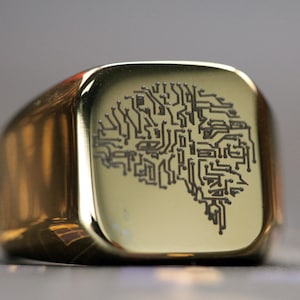 May include: A gold signet ring with a square face. The ring's face displays a black circuit board design in the shape of a brain. The ring has a polished, reflective surface. The ring is a statement piece.
