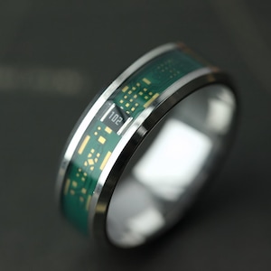 Puede incluir: Un anillo plateado con un diseño de placa de circuito verde. El anillo tiene una banda de metal pulido y una incrustación verde con detalles dorados. El número "102" es visible en la incrustación verde.