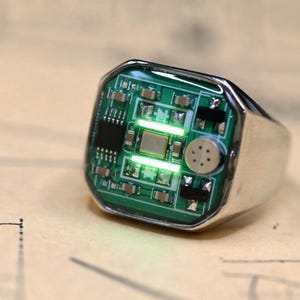 May include: A silver-toned ring featuring a rectangular face embedded with a green circuit board. The electronic component display includes small metallic chips, resistors, and two horizontal light-emitting strips that glow bright green.