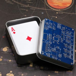 May include: A silver metal tin containing a deck of playing cards with the ace of diamonds visible on top. The lid of the tin is detached and features a blue printed circuit board design with various electronic components and silver metal screws.