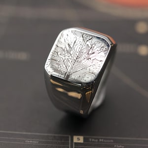 Puede incluir: Un anillo plateado con una cara rectangular. La cara presenta un diseño detallado de placa de circuito grabado en el metal. El anillo tiene una superficie pulida y reflectante, y el diseño es de un color contrastante.