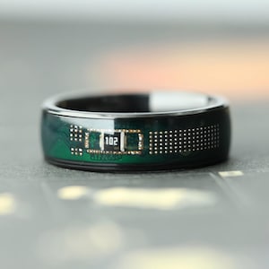 May include: A close-up of a black ring with a green circuit board design. The ring has the number "102" printed on it. The ring is a modern, tech-inspired accessory.