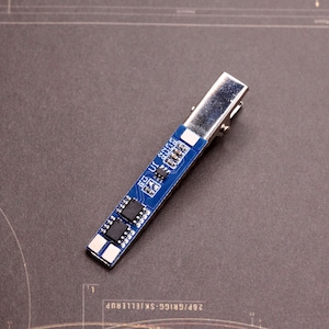 May include: A hair clip featuring a rectangular blue printed circuit board attached to a silver-toned metal alligator clip. The circuit board displays electronic components including black integrated circuits, resistors, and white labels U1, C1, R1, C2, and R3.