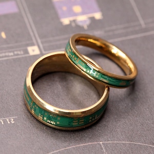 May include: Two gold-toned rings featuring a central inlay of green circuit board patterns. The rings have a polished metallic finish and a smooth, rounded band design. The green inlay displays intricate gold-coloured electronic traces and components.
