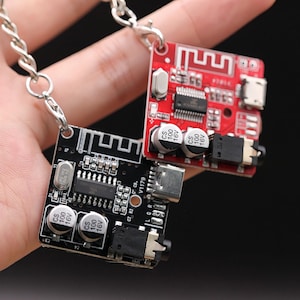 May include: Two small circuit boards, one black and one red, are attached to silver keychains. The boards have various electronic components, including capacitors and chips. The red board has a Bluetooth symbol. The boards are held up against a blurred background.