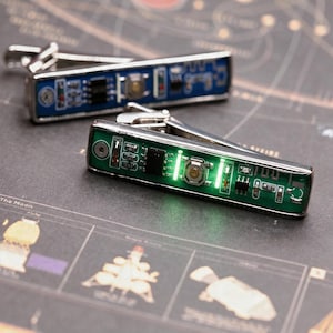 Peut inclure: Une paire de boutons de manchette rectangulaires dotés de circuits imprimés. L'un possède un circuit bleu, l'autre un circuit vert avec deux lumières LED vertes brillantes allumées de chaque côté d'un bouton métallique central.